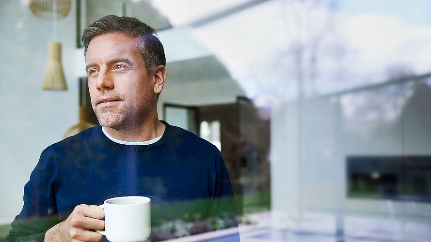 Mann mit Kaffetasse träumt und schaut dabei aus dem Fenster